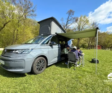 Sivý VW California T7 Beach Camper s otvorenou strechou a vysunutou markízou zaparkovaný na tráve, pri stole sedia ľudia.