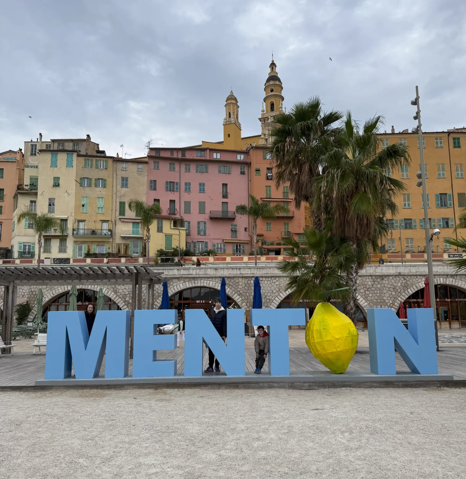 Nápis Menton na promenáde pri mori s pastelovými domami v pozadí vo Francúzsku